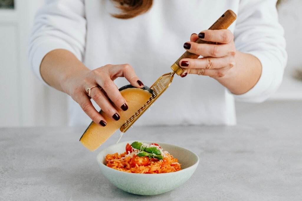 A woman with manicured nails grates parmesan over a tomato risotto, emphasizing culinary creativity.